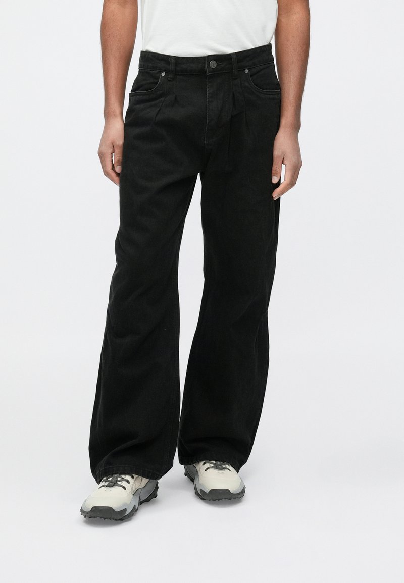 Czarne szerokie jeansy o gładkiej fakturze, z przednimi kieszeniami, zapięciem na guzik i luźnym krojem, zestawione z szaro-białymi trampkami.