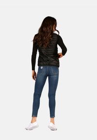 Gilet en duvet noir avec un design matelassé, porté par-dessus une chemise noire à manches longues, associé à un jean skinny bleu et des chaussures blanches.