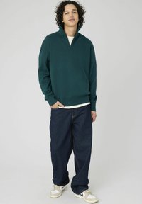 Jeune homme aux cheveux bouclés portant un pull à col zippé vert foncé, un jean ample foncé et des baskets blanches, se tenant avec une main dans sa poche.