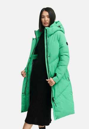 Doudoune verte avec capuche, dotée d'une fermeture éclair, d'un design matelassé et de poches latérales. Superposée sur une robe noire côtelée.