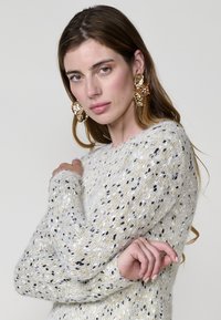 Gestufter, strukturierter Pullover in Weiß mit bunten Flecken. Weiches Material mit langen Ärmeln, ergänzt durch auffällige goldene Ohrringe.