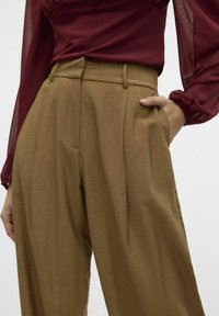 Pantalons plissés marron au tissu lisse, taille haute et poches latérales. Associés à un haut transparent à manches longues bordeaux.