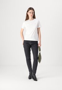 T-shirt bianca in cotone, design aderente, abbinata a jeans neri slim. Il modello tiene una borsa verde e indossa stivaletti neri alla caviglia. Linee semplici e pulite.
