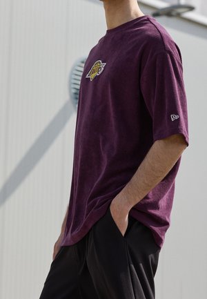Person trägt ein lila Los Angeles Lakers T-Shirt mit kleinem Logo am Ärmel, steht draußen mit den Händen in den schwarzen Hosentaschen.