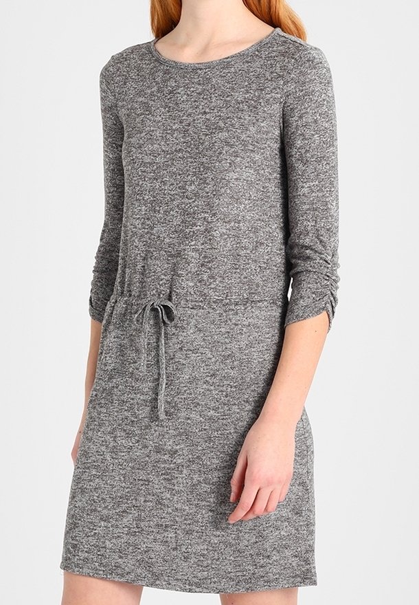 Femme portant une robe en maille gris chiné à manches longues avec taille à cordon, debout devant un fond uni clair.