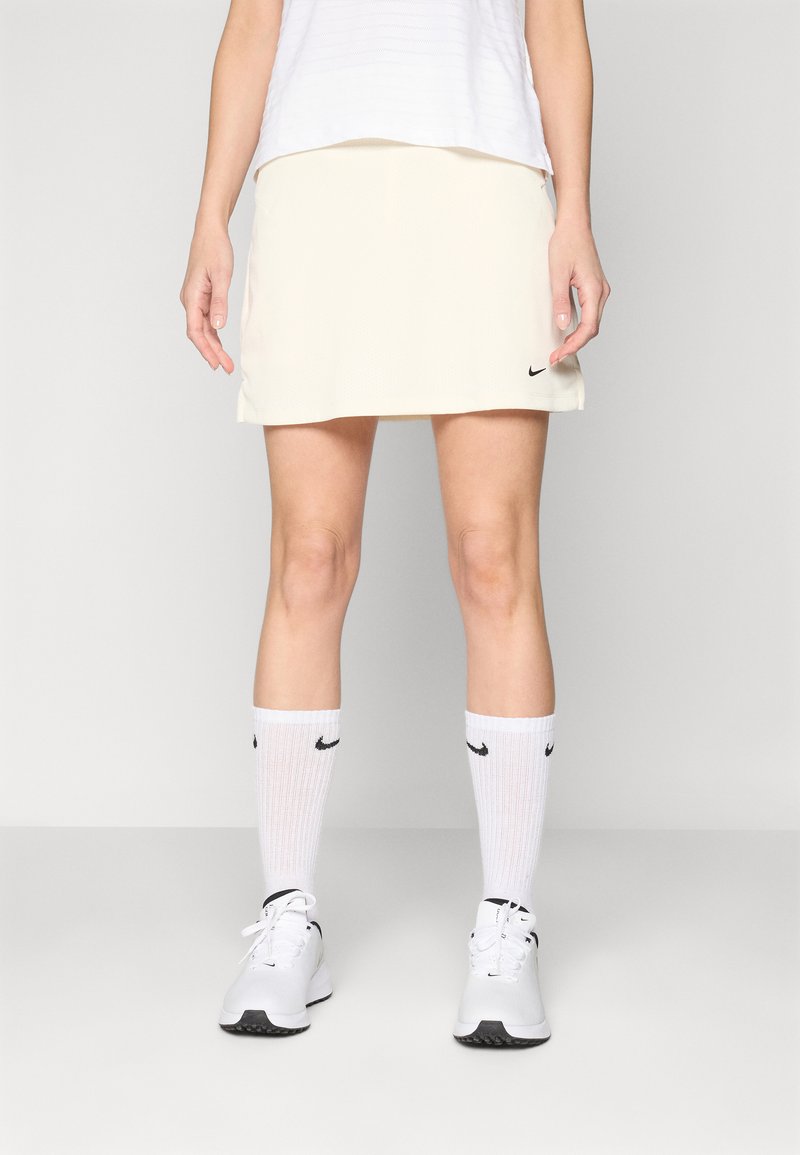 Sportovní sukně Nike v světlé krémové barvě, spárovaná s bílými ponožkami s černými logy Nike a bílými teniskami. Hladká tkanina a jednoduchý design.