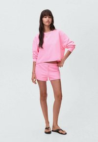 Roza skrajni pulover in usklajene roza hlače, oboje iz mehke tkanine z elastičnim pasom. Model nosi črne japanke.