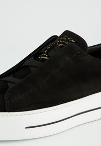 Schwarze Wildleder-Sneaker mit weißen Gummisohlen, schwarzen Schnürsenkeln und goldener Schrift auf der Zunge, präsentiert auf einem schlichten hellen Hintergrund.