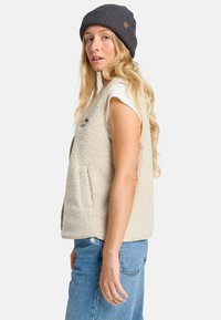Beige fuzzy fleece vest with wide arm openings, side pockets, and a round neckline. Paired with a dark gray knitted beanie.