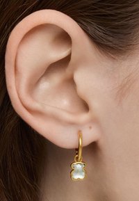 Goldene Creolen mit einem transparenten, klobigen Charme mit facettiertem Oberflächenmuster. Die Ohrringe hängen unterhalb des Ohrläppchens und betonen die Details.