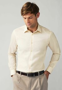 Chemise beige ajustée à manches longues, avec un devant boutonné et un col. Portée avec un pantalon marron clair et une ceinture marron foncé.