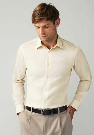 Beige slim-fit särk pika varrukaga, nupukinnise ja kraega. Kanda heledate pruunide pükstega ja tumeda pruuni vöödiga.