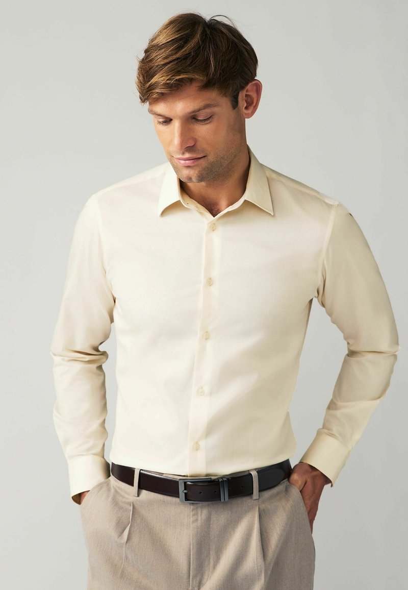 Chemise beige ajustée à manches longues, avec un devant boutonné et un col. Portée avec un pantalon marron clair et une ceinture marron foncé.