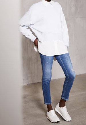 Personne portant un sweat-shirt blanc oversize superposé sur une chemise blanche, un jean skinny bleu déchiré et des baskets blanches, debout avec une main sur la hanche.