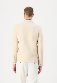 Beige zakó hosszú ujjal és gombos mandzsettával, egyszerű designnal és sima textúrával. Minimalista stílus, látható minták nélkül.