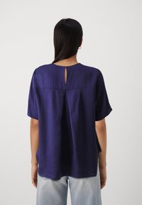 Marineblaue Kurzarmbluse mit rundem Halsausschnitt, ausgestattet mit einer Schlüsselöffnung auf der Rückseite und einem lässigen Schnitt. Glatte Stofftextur.