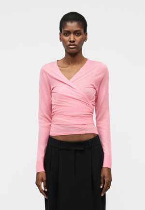 Žena s tmavou pokožkou, ktorá má na sebe svetloružový zavinovací top s dlhými rukávmi a čierne nohavice s vysokým pásom, stojaca pred jednoduchým pozadím.