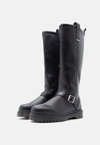 Et par høye sorte lærstøvler med spennefester og sydde detaljer, med tykke såler med mønster på hvit bakgrunn.