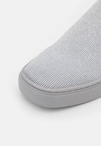 Gros plan d’un bout de chaussure de sport en tissu texturé gris clair avec une semelle en caoutchouc lisse, photographié sur fond blanc.