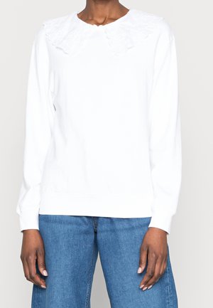Personne portant un haut blanc à manches longues avec un large col à volants et un jean bleu en denim sur un fond clair uni.