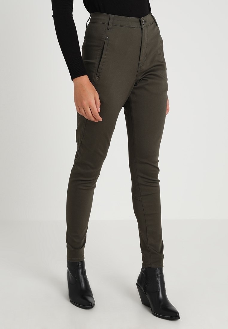 Femme portant un pantalon slim fit vert olive avec fermeture par bouton et zip, poches avant, associé à des bottines noires et un haut noir à manches longues.