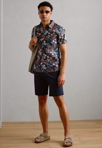 Blomstret kortærmet skjorte i marineblå med lyse farver, parret med mørke shorts og lyse sandaler. Holder en beige taske.