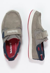 Zapatos de gamuza gris y tela con una correa de Velcro, media suela blanca, detalles en rojo y el logo de Timberland en el lateral. Diseño cómodo para uso casual.