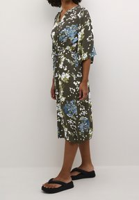 Blumenkleid in Olivgrün mit blauen und weißen Blumen, kurzen Ärmeln, V-Ausschnitt und einer Taillenschnürung. Kombiniert mit schwarzen Slip-On-Sandalen.