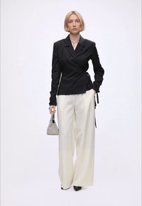 Femme aux cheveux blonds courts portant un blazer noir croisé, un pantalon blanc large, des chaussures noires et tenant un petit sac à main texturé sur un fond uni.