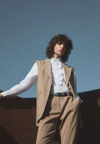 Beige gestreifter Weste und passende Hose über einem weißen, gerüschten Hemd, kombiniert mit einem schwarzen Gürtel. Der Stoff wirkt glatt vor einem blauen Himmel.