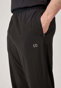 Czarne sportowe spodnie z elastycznym paskiem w talii, boczną kieszenią oraz małym szarym logo z przodu. Gładka, lekka tekstura materiału.