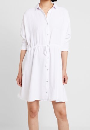 Kobieta ubrana w białą sukienkę zapinaną na guziki, z kołnierzykiem, wiązaną w talii sznurkiem i rękawami do połowy łokcia na tle jednolitego jasnoszarego tła.