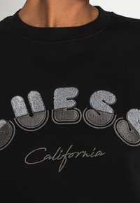 Černá mikina s kulatým, metalickým stříbrným vyšitým nápisem "Guess California." Výstřih je ribbený a těsně padnoucí.