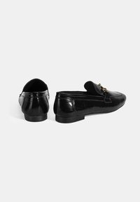 Zwarte leren loafers met een glanzende afwerking, voorzien van een decoratieve gouden hardware accent en een lage, platte hak. Geverfde textuur op de zijkanten.