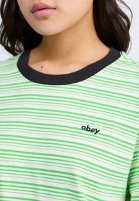 Žalias ir baltas dryžuotas marškinėliai su juodu gipiūru aplink kaklą ir mažu "obey" logotipu kairėje krūtinės pusėje. Minkšta, lengva medžiaga.