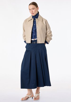 Femme portant une veste courte beige sur une chemise boutonnée bleu marine, une longue jupe plissée bleu marine, et des sandales beige à petit talon, debout les mains derrière le dos.