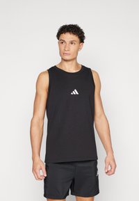 Camisola desportiva preta sem mangas feita de uma mistura de algodão, com um pequeno logótipo branco da Adidas no peito, acompanhada de calções escuros.