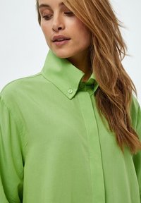 Camicia verde con colletto, realizzata in tessuto morbido, con vestibilità rilassata e dettagli di cucitura ben visibili.