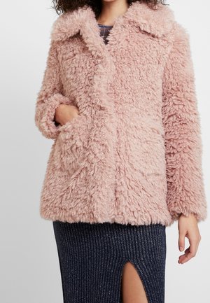 Nainen, jolla on päällään pörröinen vaaleanpunainen tekoturkistakki suurella kauluksella, käsi taskussa, yhdistetty tumman ribbineuloksisen hameen kanssa, jossa on etuslitsi.