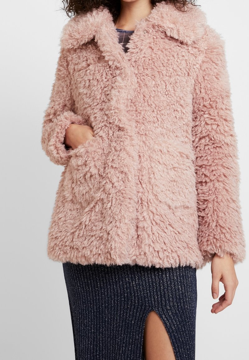 Femme portant un manteau en fausse fourrure rose pâle et duveteux avec un grand col, main dans la poche, associée à une jupe côtelée foncée avec une fente sur le devant.