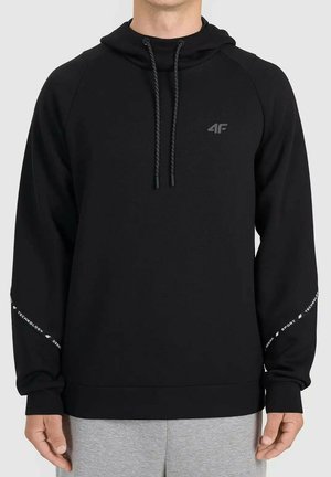 Mężczyzna w czarnej bluzie z kapturem z logo "4F" na klatce piersiowej i białym tekstem na rękawach, w połączeniu z szarymi spodniami.