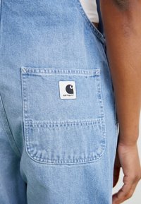 Blå denimslitoveraller har en baklomme med en hvit etikett som viser en stilisert logo. Stoffet er mykt med en falmet finish.