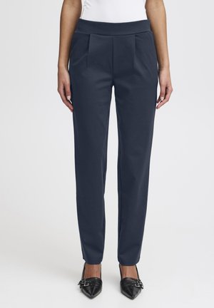 Pantaloni navy sartoriali con una texture liscia, dotati di una cintura larga e pinces anteriori, abbinati a scarpe nere con punta affusolata.