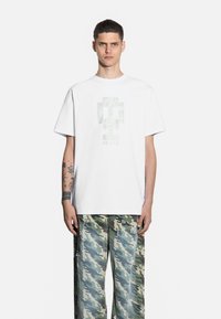 Biała bawełniana koszulka z teksturowanym wzorem graficznym na środku. W komplecie z luźnymi spodniami w zielony kamuflaż, z postrzępieniami.