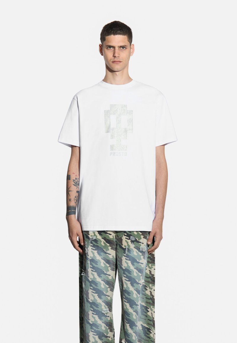 Biała bawełniana koszulka z teksturowanym wzorem graficznym na środku. W komplecie z luźnymi spodniami w zielony kamuflaż, z postrzępieniami.