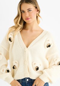 Cream-colored knitted cardigan with a V-neck, large buttons, and textured bear motifs on sleeves. Soft, chunky yarn provides a cozy appearance.