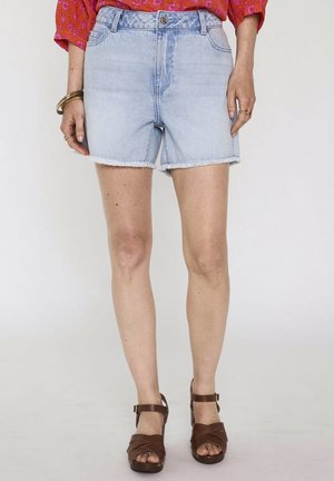 Short en denim bleu clair avec ourlets effilochés, porté avec des sandales marron à talons et un haut rouge à motifs, visible de la mi-cuisse à la taille.