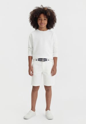 Παιδί με σγουρά μαλλιά που φοράει λευκό μακρυμάνικο πουκάμισο, λευκά σορτς με ριγωτή ζώνη και λευκά αθλητικά παπούτσια, στέκεται μπροστά σε απλό φόντο.