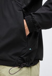 Persoon draagt een zwart jack met elastische manchetten en hand in de zak, beige broek zichtbaar, klein Zweeds vlaggetje bij de zoom.