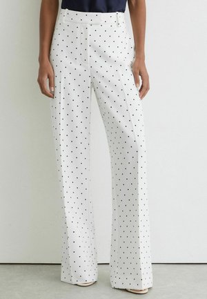 Witte broek met wijde pijpen en zwarte stippen, gedragen door een persoon die voor een effen witte achtergrond staat.
